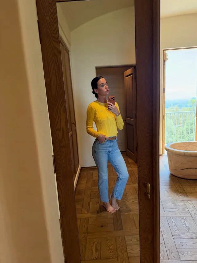 Katy Perry standing barefoot indoors in jeans and a yellow top.