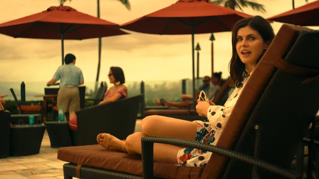 Alexandra Daddario reclining barefoot on an outdoor chair beneath patio umbrellas.