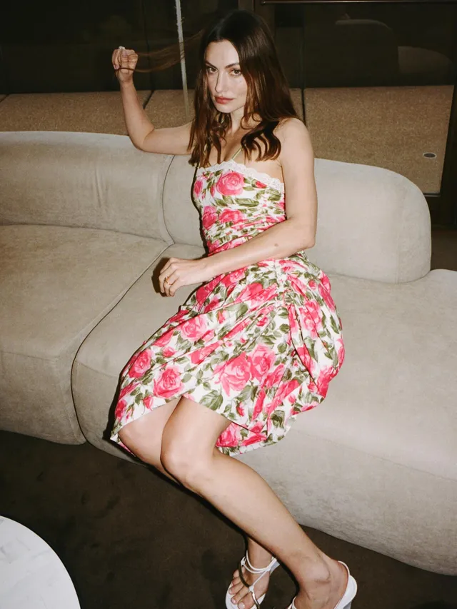 Phoebe Tonkin sitting on a sofa in a floral dress and heels.