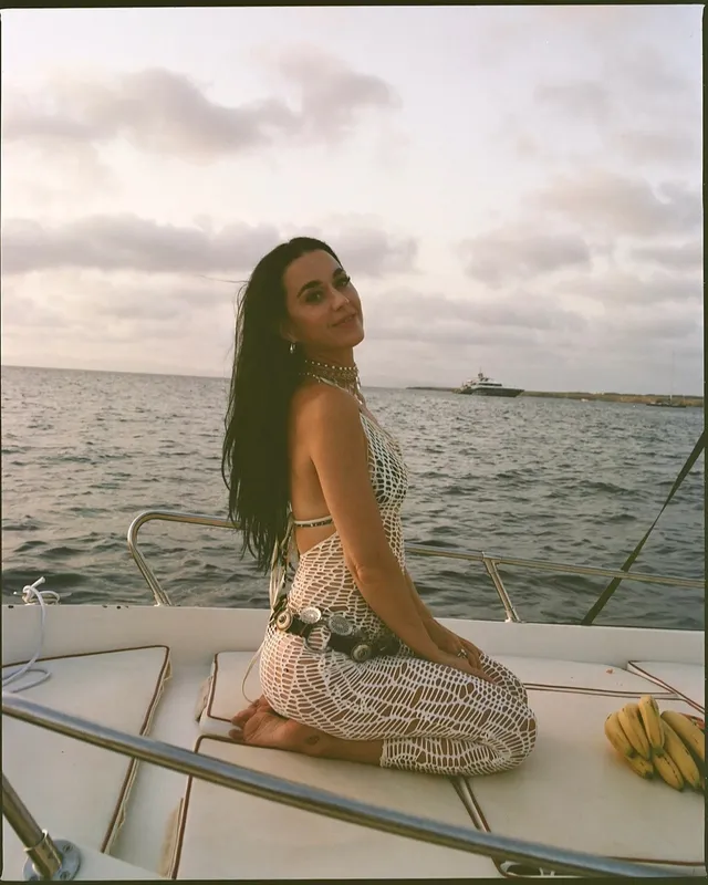 Katy Perry kneeling barefoot on a boat deck beside the sea.