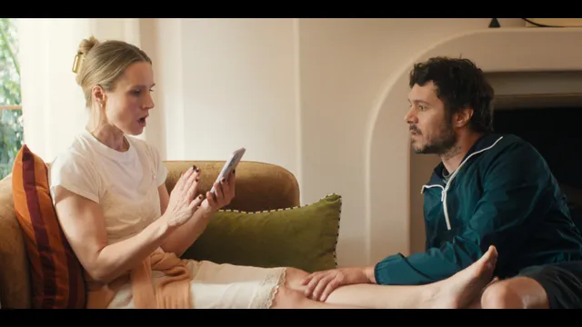 Kristen Bell sitting on a sofa with bare feet beside another person.