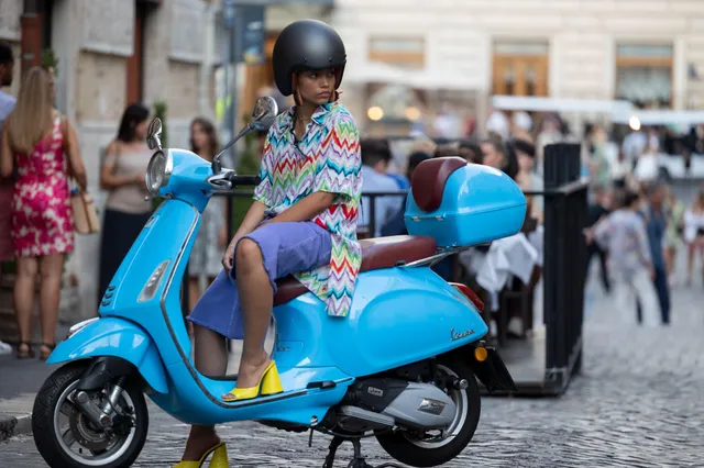 Whitney Peak sitting on a blue scooter in a patterned outfit and bright yellow heels.