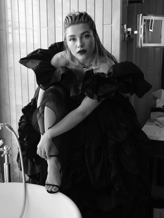 Florence Pugh sitting in a black dress and heels in a monochrome portrait.