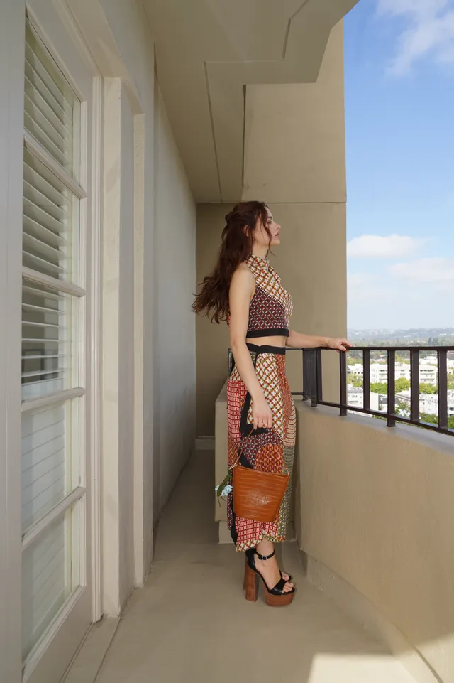 Joey King standing on a balcony in a patterned outfit and platform heels.