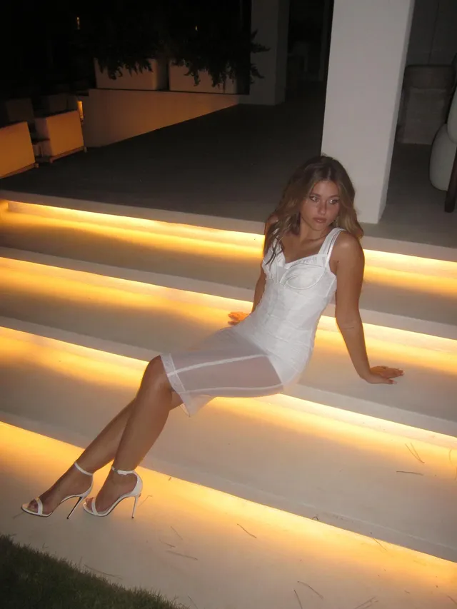 Nicole Wallace sitting on illuminated stairs in a short white dress and heels.