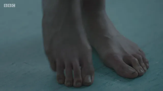 Jodie Comer standing barefoot in a dimly lit screen scene.