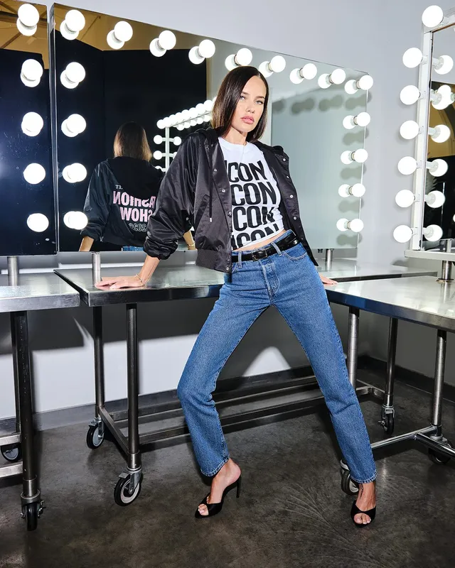 Adriana Lima standing beside a dressing-room mirror in jeans and black heels.