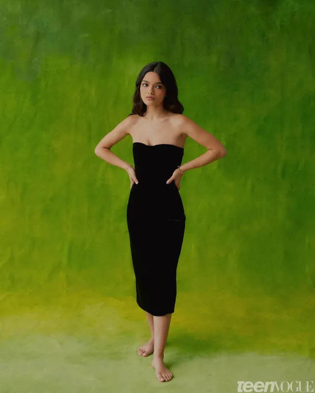 Rachel Zegler posing barefoot in a black dress against a green backdrop.