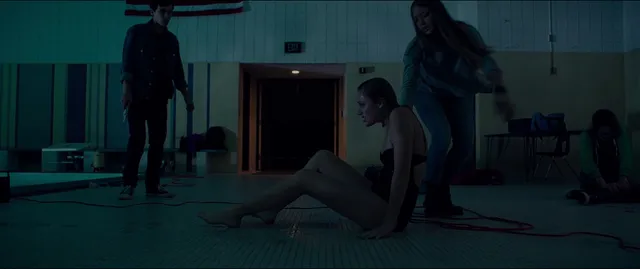 Maika Monroe sitting barefoot on a floor in a dimly lit scene.