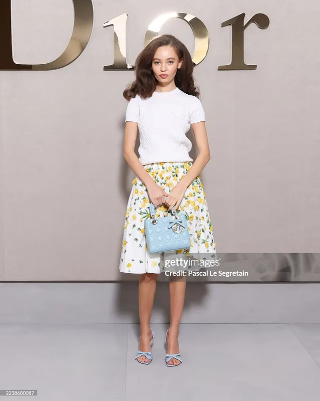 Nico Parker standing at a fashion event in a patterned skirt and pale heels.