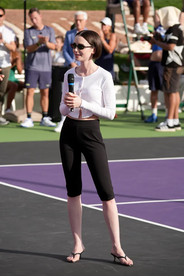 Lily Collins standing on a tennis court in cropped trousers and flip-flops.