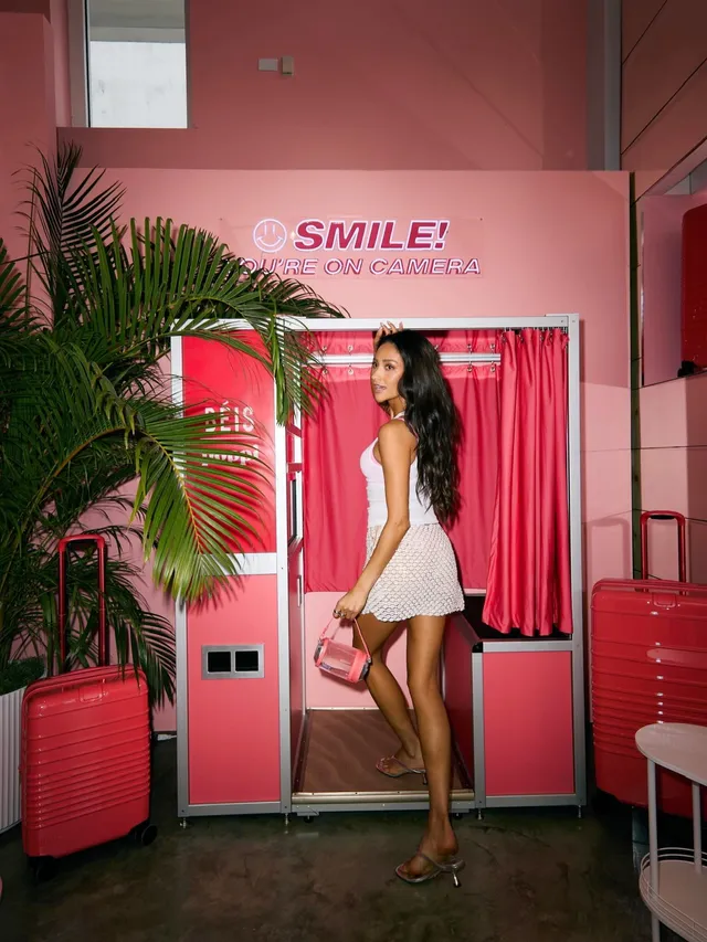 Shay Mitchell posing beside a pink luggage display in a short white dress.