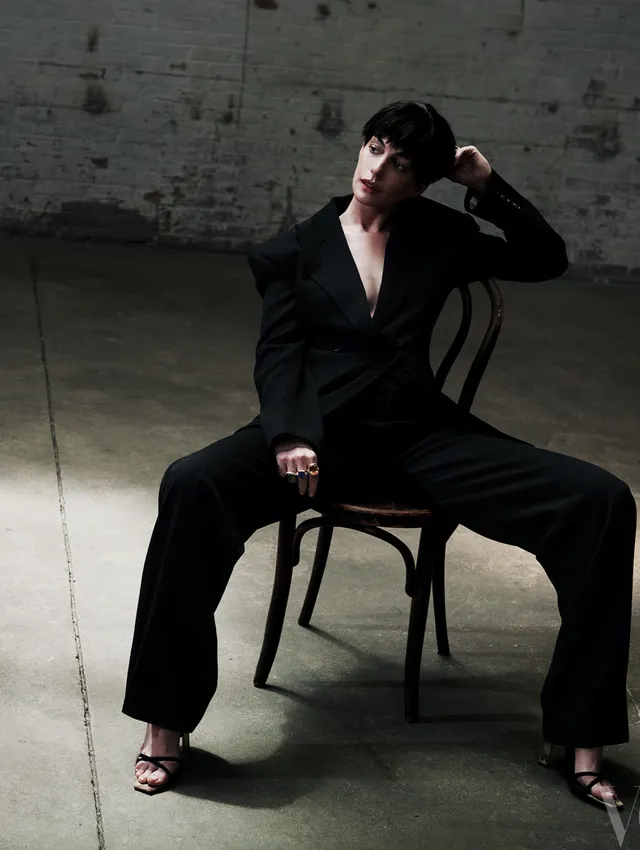 Anne Hathaway sitting on a chair in a black suit and heels.