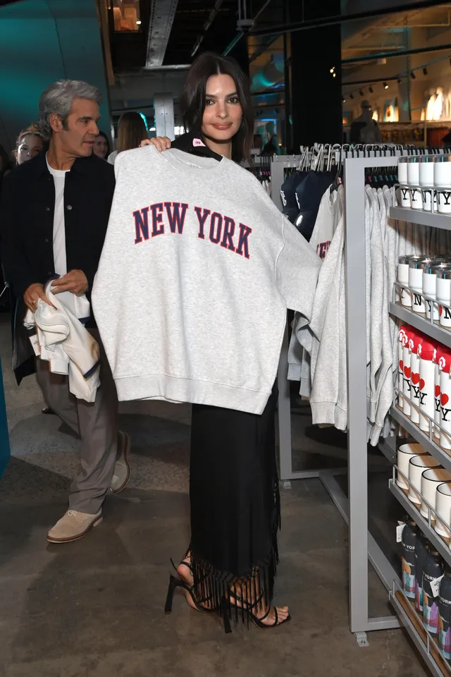 Emily Ratajkowski holding a white sweater while wearing a black skirt and heels.