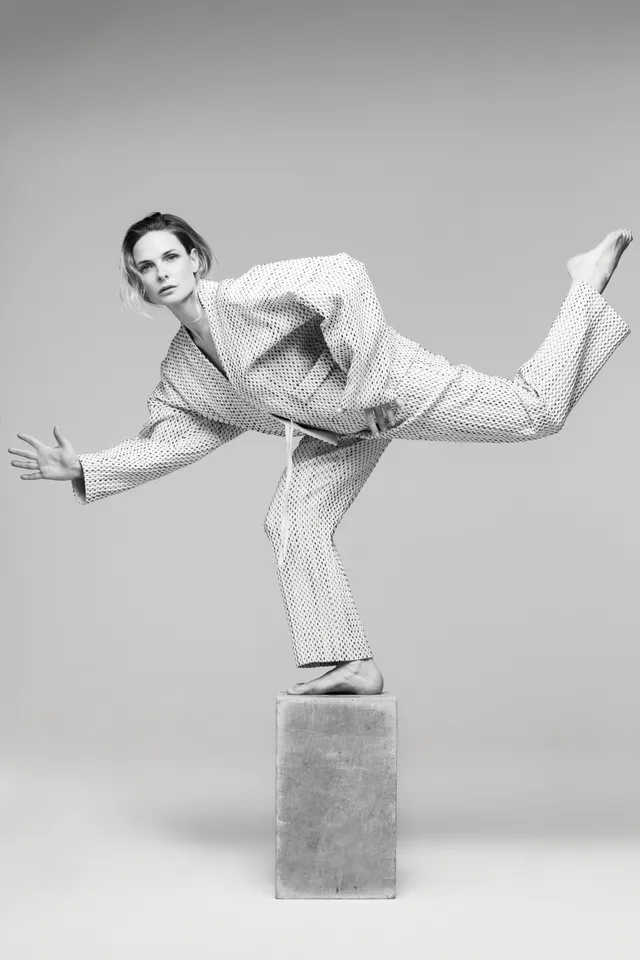 Rebecca Ferguson balancing in a tailored suit and heels in a monochrome studio portrait.