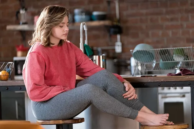 Chloë Grace Moretz sitting barefoot on a kitchen counter in side view.