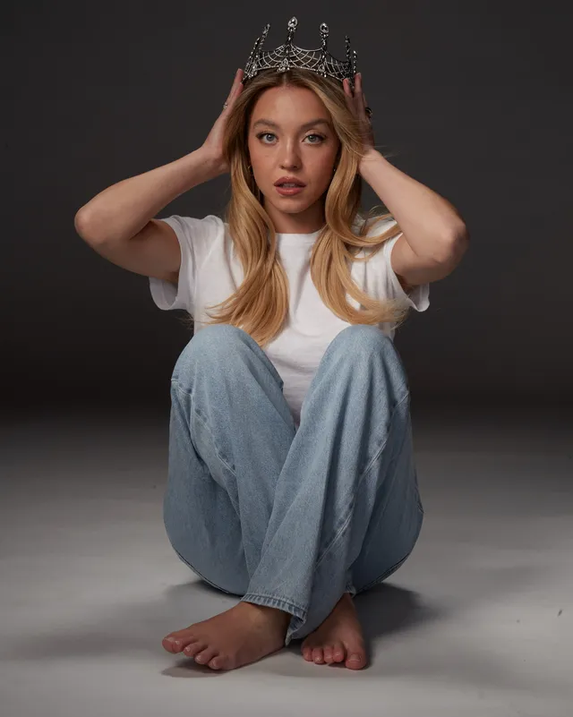 Sydney Sweeney sitting against a dark backdrop in a white T-shirt, jeans and a decorative crown.