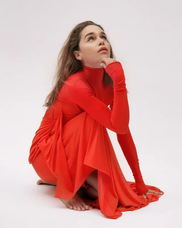 Emilia Clarke crouching barefoot in a long red dress.