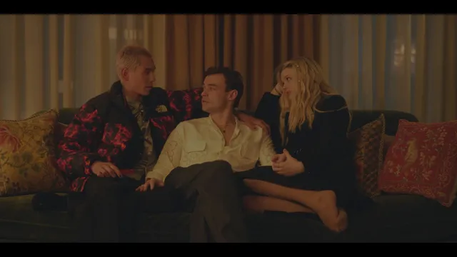 Emily Alyn Lind sitting barefoot with two men on a sofa in a scene.
