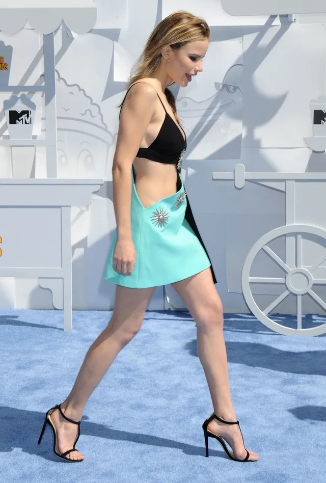 Halston Sage walking on a carpet in a black top, turquoise skirt and black heels.