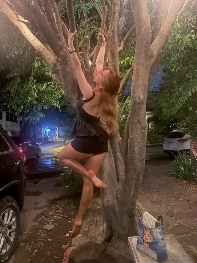 Addison Rae posing barefoot beside a tree at night.