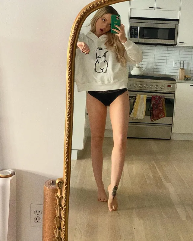 Dove Cameron posing barefoot beside a kitchen mirror.