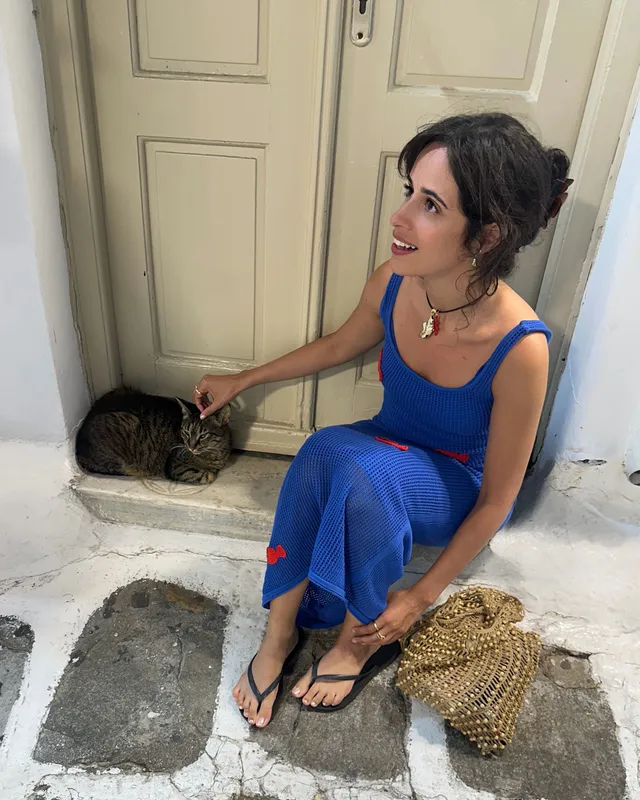 Camila Cabello sitting beside a doorway in a blue dress and flat sandals.