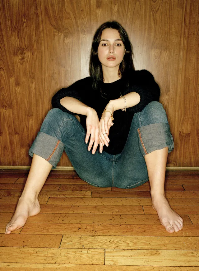 Keira Knightley seated against a wooden wall in jeans and a black top.