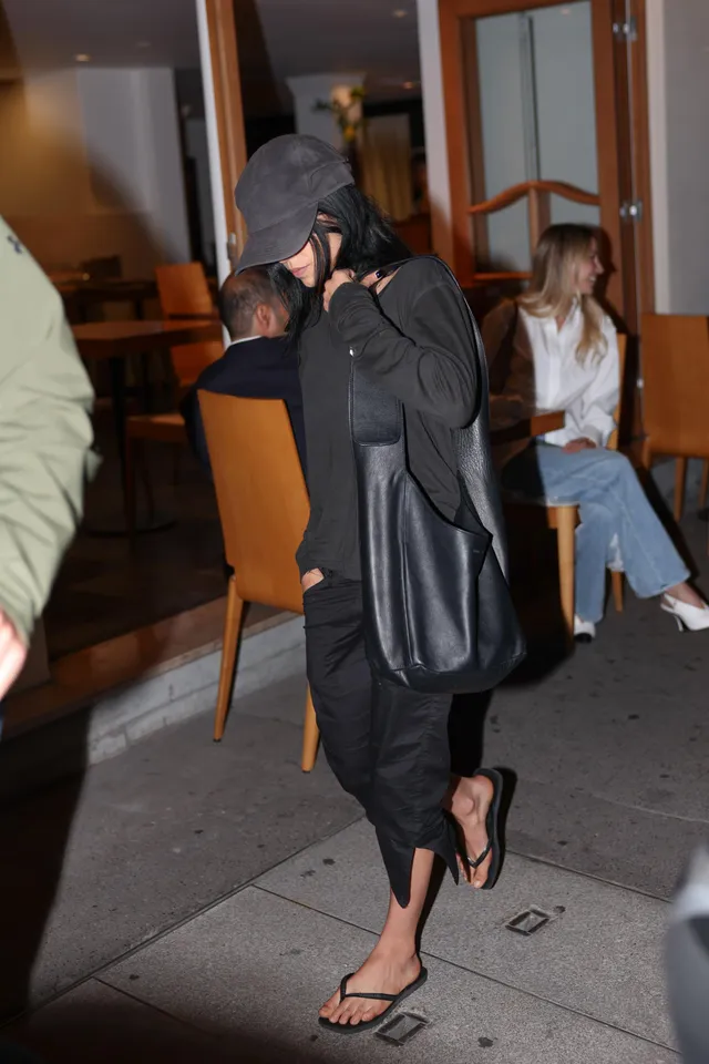 Jenna Ortega walking outside in a black outfit, cap and flat sandals.