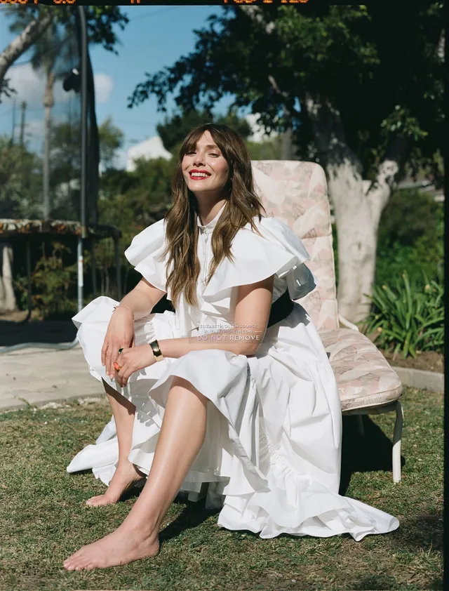 Elizabeth Olsen sitting barefoot outdoors in a white dress.
