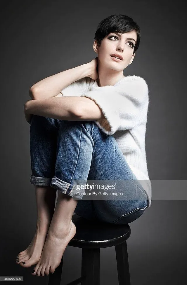 Anne Hathaway sitting barefoot on a stool in jeans.