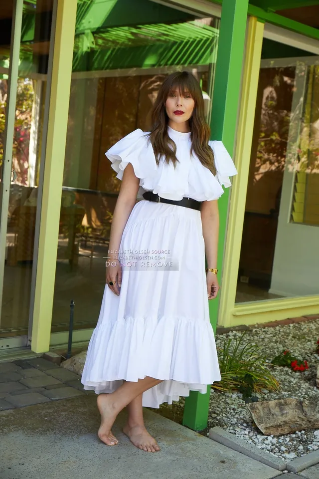 Elizabeth Olsen standing barefoot in a white dress by green doors.