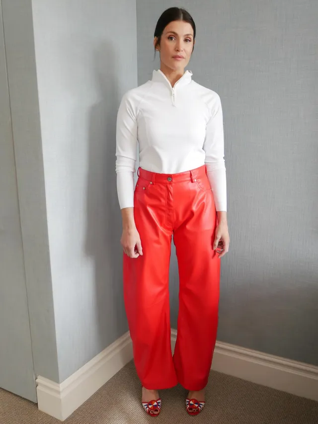 Gemma Arterton standing beside a doorway in red trousers.