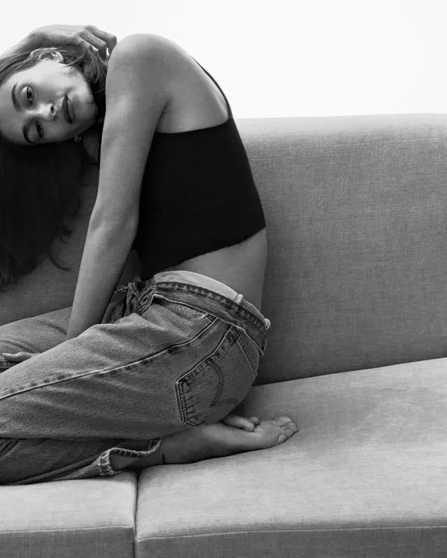 Hailey Bieber sitting barefoot on a sofa in a monochrome portrait.