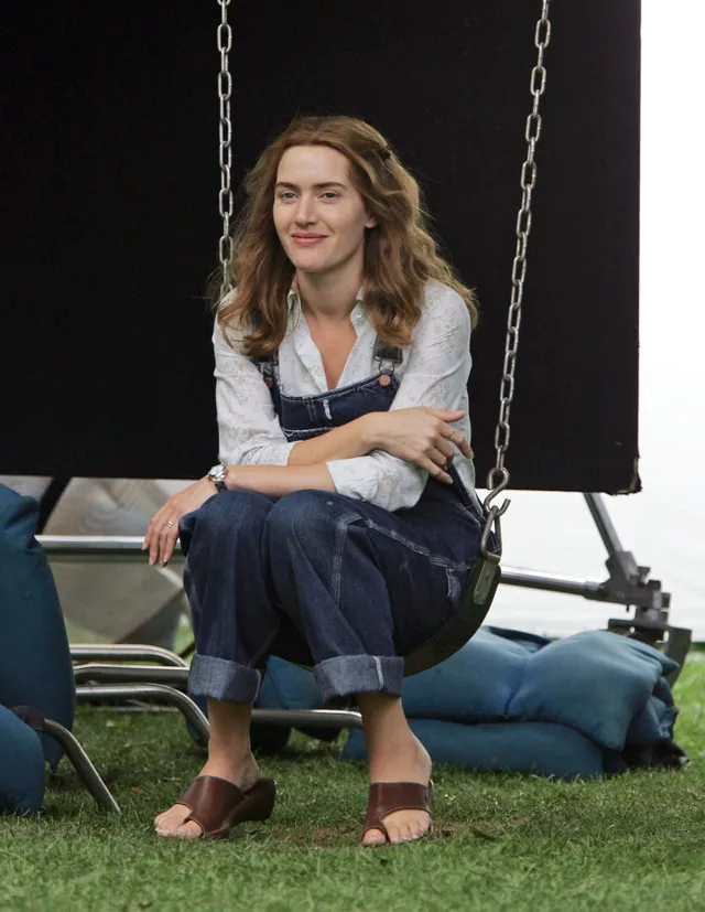 Kate Winslet sitting outdoors in denim overalls and sandals.