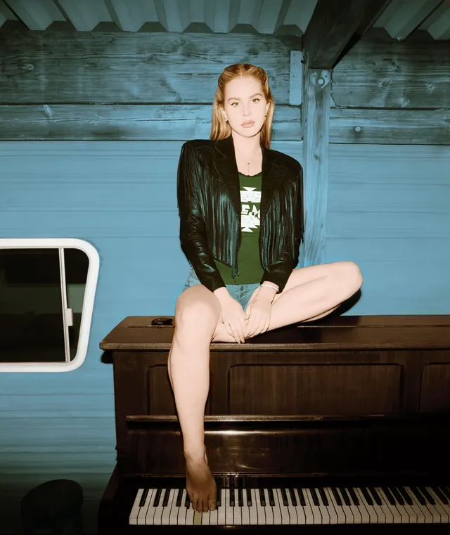 Lana Del Rey sitting barefoot on a piano in a jacket and green outfit.