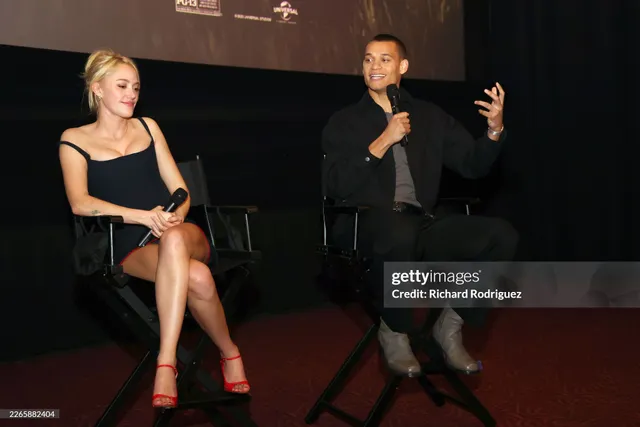 Maika Monroe sitting onstage in a black dress and red heels.