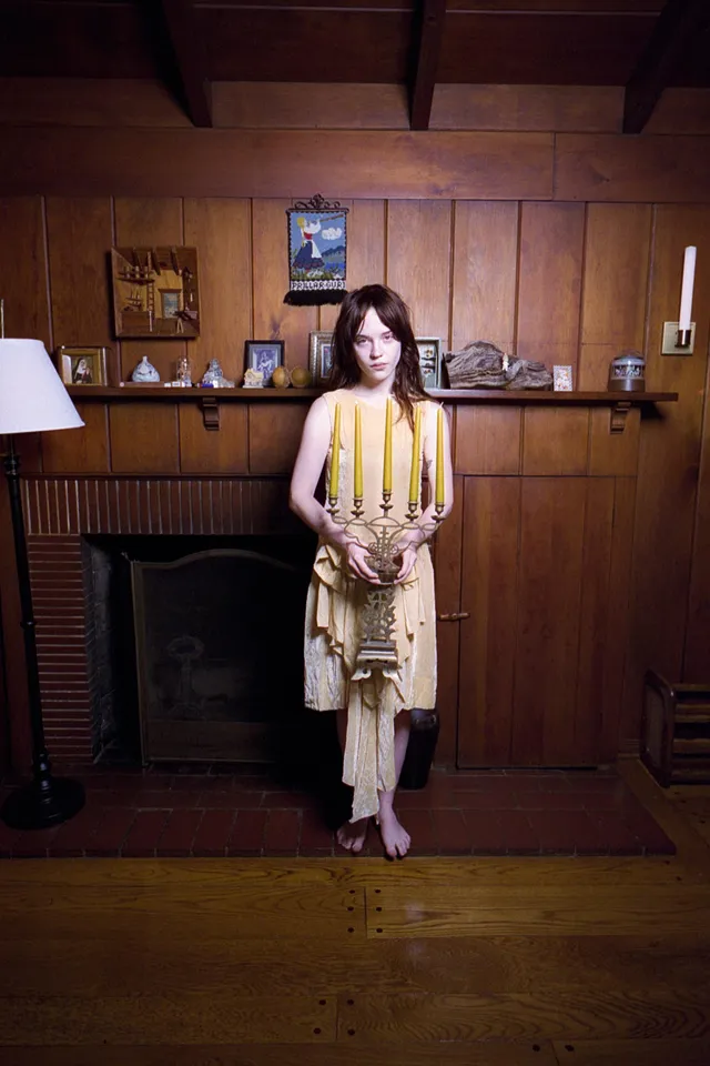 Sophie Thatcher standing barefoot beside a fireplace in a patterned dress.