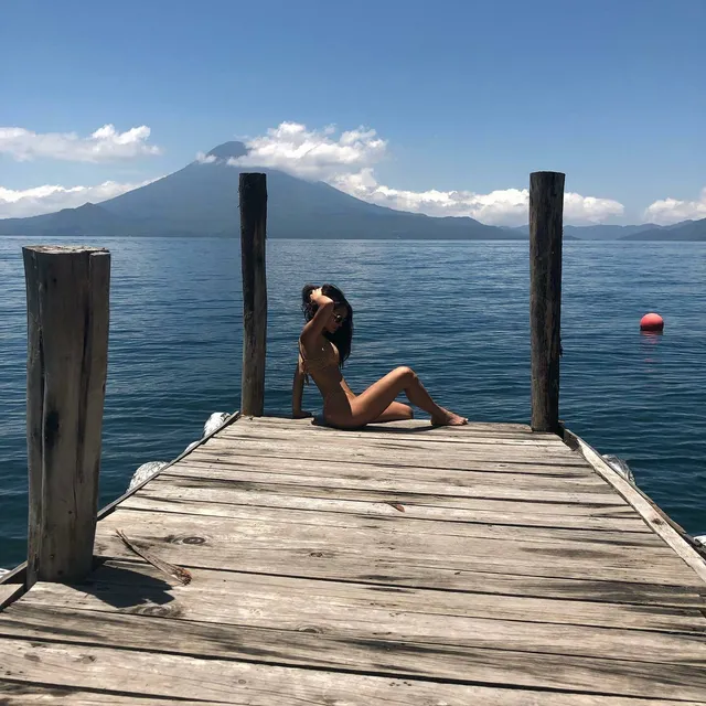 Adria Arjona sitting barefoot on a wooden jetty beside a lake.