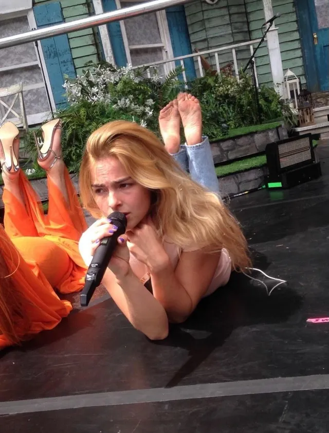 Addison Rae lying on a stage while singing with her bare feet raised.