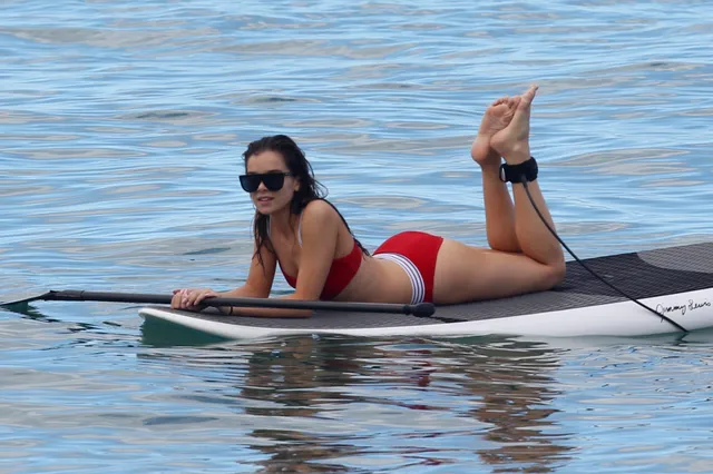 Hailee Steinfeld lying barefoot on a paddleboard on blue water.