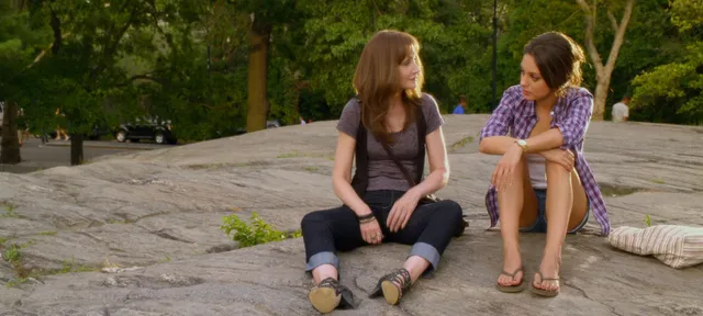 Mila Kunis sitting in a park beside another adult while wearing heeled sandals.