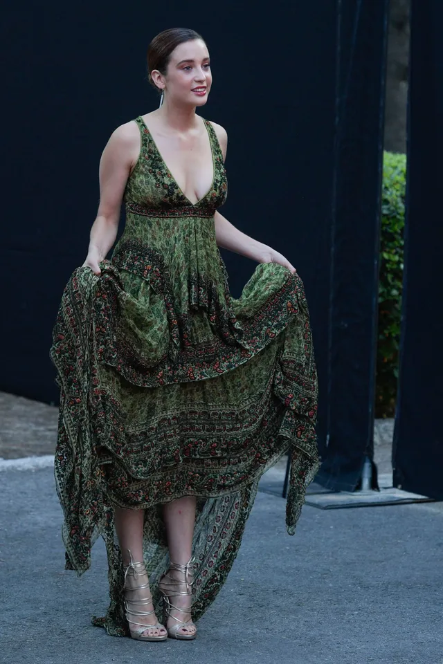 María Pedraza walking outdoors in a long green dress and metallic heels.