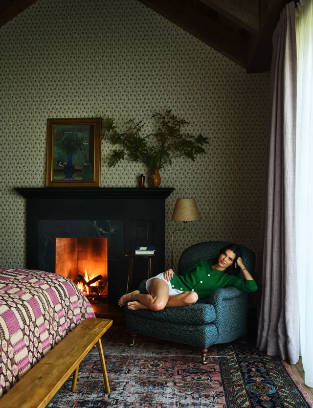Kendall Jenner reclining barefoot in a green armchair by a fireplace.