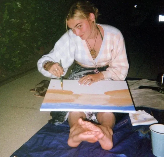 Meg Donnelly sitting barefoot on a picnic blanket while painting.