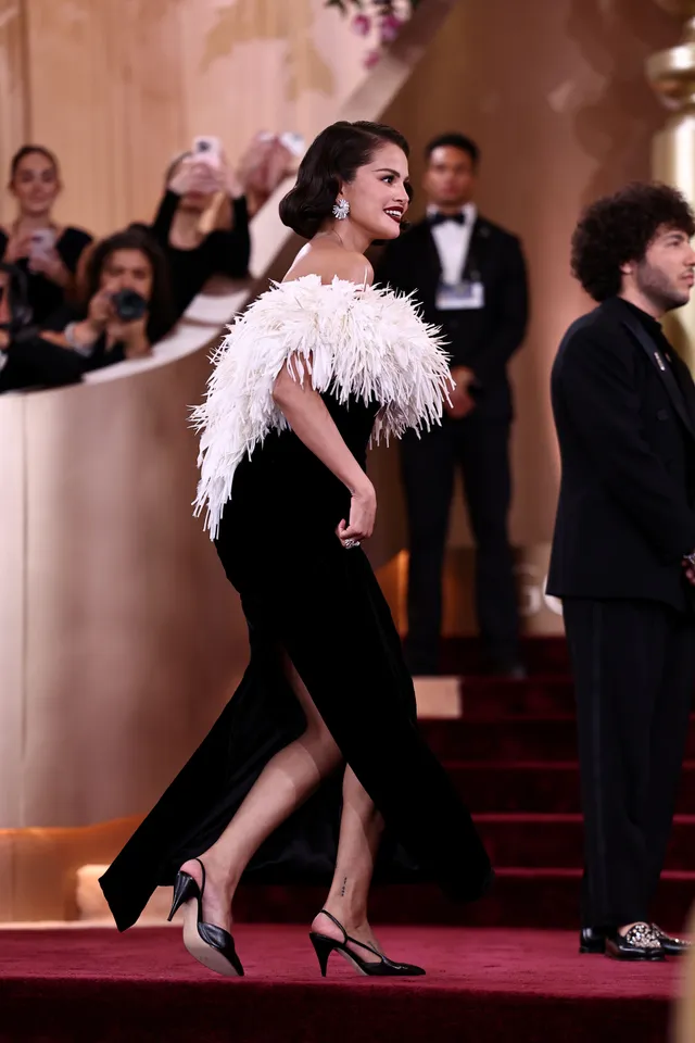 Selena Gomez walking indoors in a black dress with feather trim.