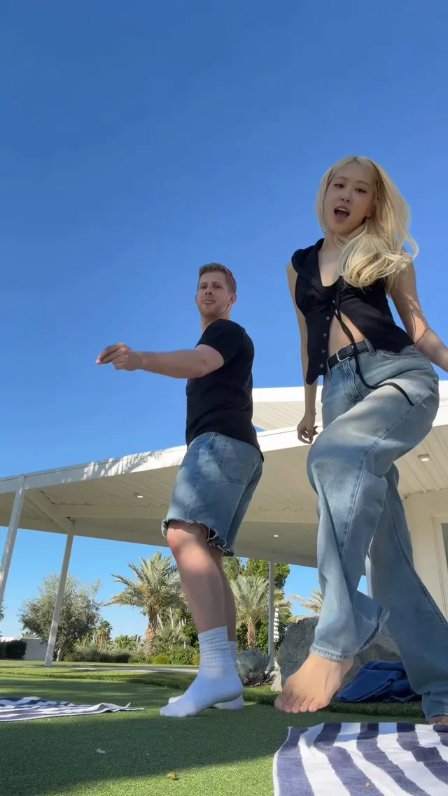 Rosé lifting one bare foot while dancing on a lawn in a dark top and jeans.