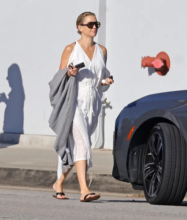Kate Hudson walking beside a car in a white dress and flat sandals.