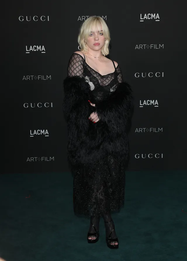 Billie Eilish standing against an event backdrop in a lace dress and heels.
