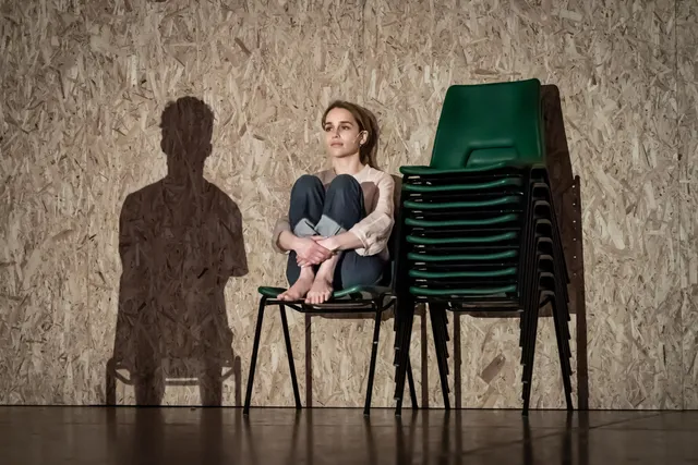 Emilia Clarke sitting barefoot beside stacked green chairs.